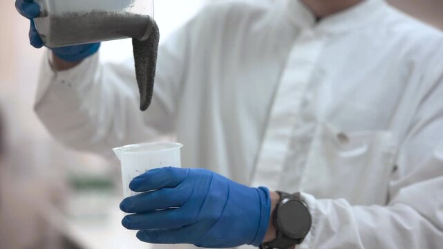 Laboratory technician pouring dark granular powder from clear plastic container into transparent measuring beaker while wearing blue nitrile gloves and white lab coat