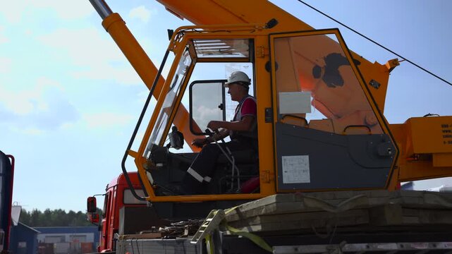 Orange mobile crane mounted on truck with seated operator in hard hat at industrial yard, side profile under bright daylight, showing boom, rigging gear, red cab, and open sky