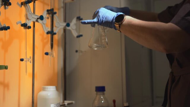 Gloved hands lifting empty Erlenmeyer flask inside laboratory fume hood beside clamp assembly and reagent bottles, with laboratory, gloved, hand, flask detail, showing erlenmeyer, glass, clamp
