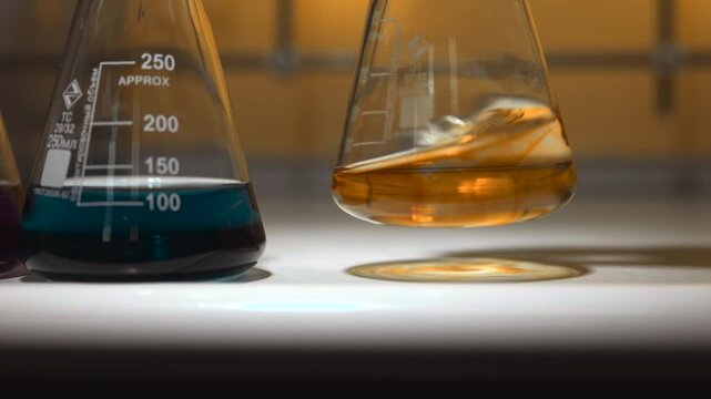 Low-angle close-up of laboratory glass flasks holding teal and amber liquids with brief lift, swirling displacement, soft reflections, warm blurred background, clean tabletop scene