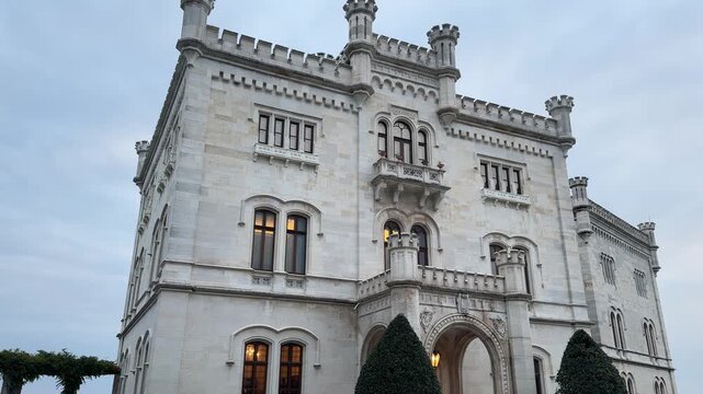 Trieste, Italy. Historic stone castle with turrets and clock tower against cloudy sky, surrounded by trees and greenery