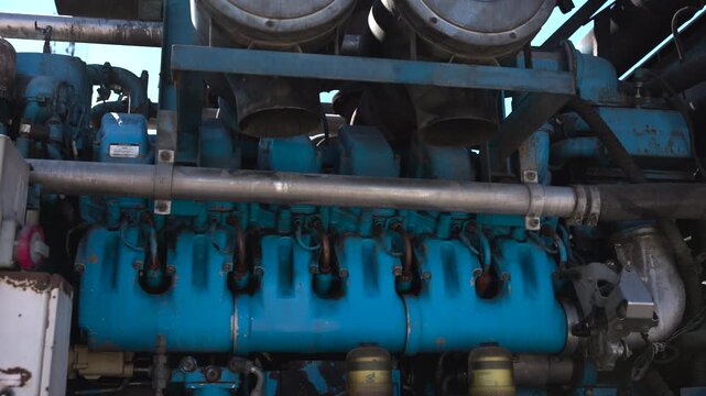 Low angle close view of teal blue industrial engine bank with manifold, piping, filters, hoses, brackets, and worn metal surfaces inside heavy machinery compartment