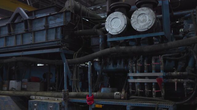 Large blue industrial machinery with dense pipe network, insulated hoses, clustered valves, heavy fittings, weathered metal surfaces, and dim warehouse interior with gritty maintenance details