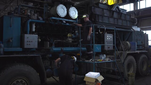 Technicians inspecting oversized diesel engine on mobile chassis inside industrial maintenance bay with raised platform, tool bench, exposed components, pipes, hoses, and safety gear