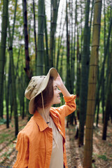 Woman in bamboo forest wearing a wide brim hat and orange shirt, smiling in profile. Portrait of nature travel and outdoor exploration with serene green background. © SHOTPRIME STUDIO