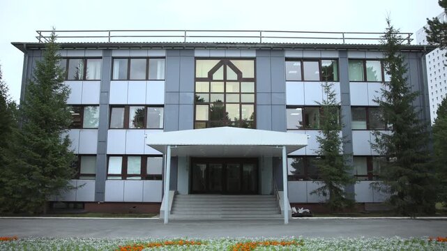Symmetrical modern institutional building with central glass entrance, covered stairway, rows of windows, gray panel facade, evergreen trees, and flower border under soft overcast daylight