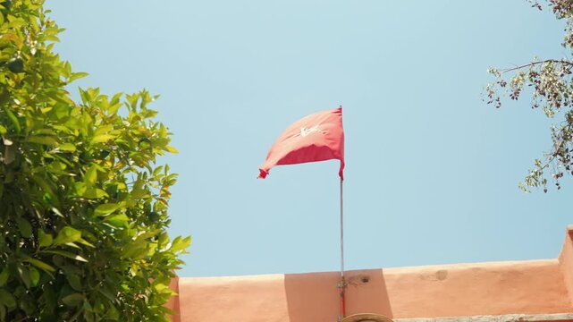 Red Flag Fluttering Above Peach Wall Against Clear Blue Sky With Green Foliage