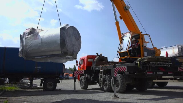 Mobile crane lifting large cylindrical metal tank in industrial yard beside trailer, with operator in cab and ground crew handling rigging, safety gear, and heavy cargo during transfer