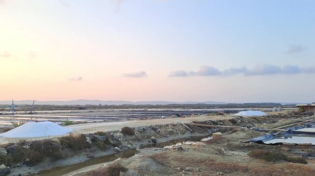 Salt farm landscape at sunset in Sumenep, Madura, Indonesia. Wide view of salt evaporation ponds with salt piles, canals and orange sky. Traditional agriculture industry.