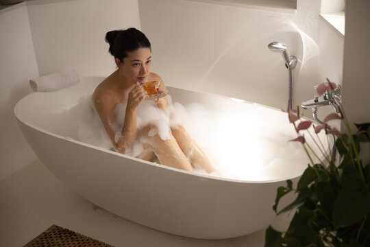 African woman sitting in freestanding oval bathtub in bathroom sipping from clear glass teacup