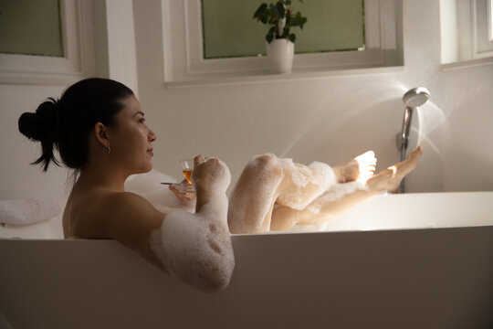 African American woman reclining in bathtub filled with soap bubbles, holding stemmed glass