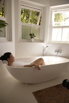 African American female reclining in oval tub with bubbles and rolled towel by windows