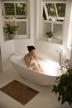 African American woman sitting in bathtub with bubbles in bathroom holding glass with amber drink