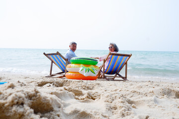 Fototapeta premium Senior couple relaxing on beach chairs under a beach umbrella with colorful inflatable swim rings by the sea on a sunny day. Peaceful summer vacation concept featuring retirement lifestyle, travel, re