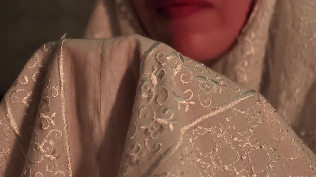 Extreme macro slow motion shot focusing on embroidered white mukena fabric covering cupped hands held up during sincere prayer.
