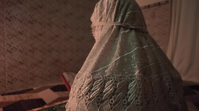 Back angle slow motion video showing a Muslim woman covered in white mukena sitting on a carpet and reading the holy Quran.
