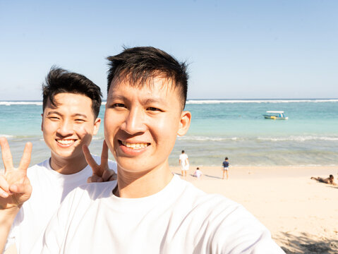 Two smiling young men pose on a sunny beach. They wear matching white t-shirts. Ocean waves roll gently  Lone boat floats far out to sea
