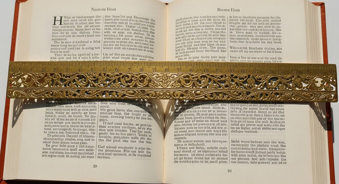 A beautifully ornate brass ruler with intricate scrollwork rests across the open pages of a hardcover book, casting delicate shadows and highlighting the printed text for focused reading
