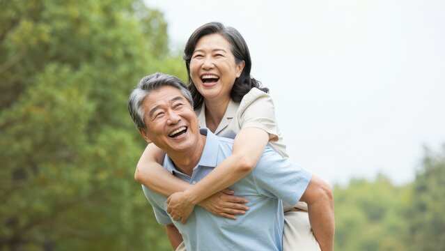 Happy senior Asian couple having fun outdoors with man giving woman a piggyback ride in a lush green park during bright daylight, expressing joy, love, and active lifestyle together