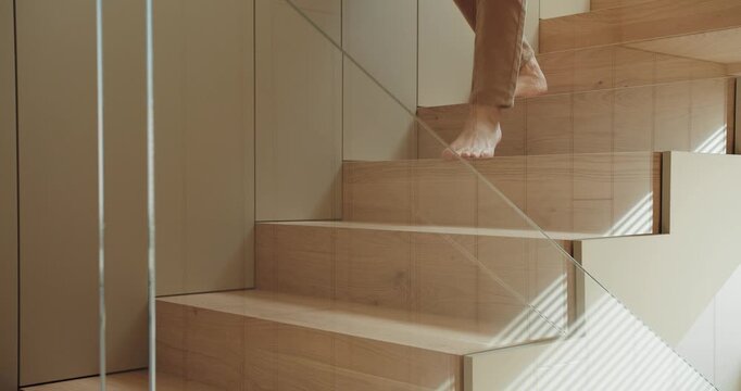 A person climbs a modern wooden staircase, showcasing bare feet and contemporary architecture with natural lighting. Close-Up of Bare Feet Walking Up Wooden Staircase in Modern Home