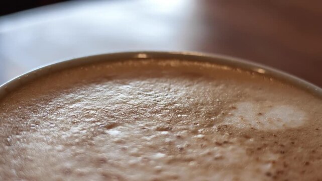 Slow motion close up shot cropped on the top rim of a pink cappuccino cup, highlighting the light foam pattern and sprinkled cinnamon on a warm wooden background.
