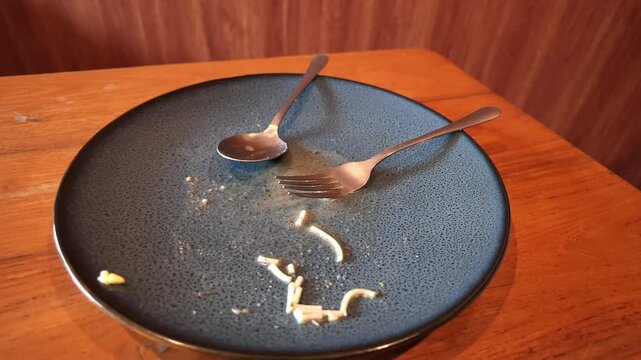 Slow motion top high angle camera shot showing an empty dark blue plate with leftover crumbs and used utensils on a wooden table.
