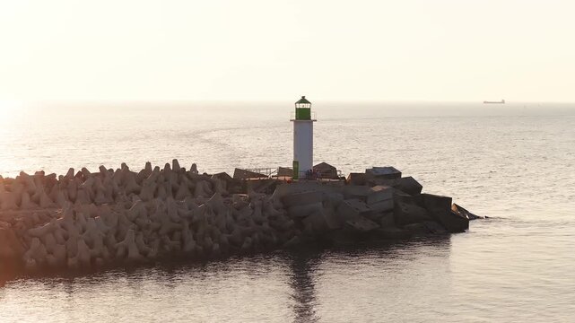 Aerial view shows a white lighthouse on tetrapods at Ventspils port, Latvia. Golden hour backlights calm Baltic Sea, long shadows, subtle waves, and a slow freighter.