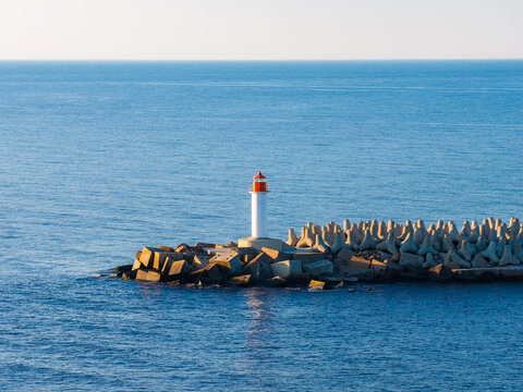 Aerial view shows the Ventspils harbor lighthouse in Latvia on tetrapod breakwater, lit by warm low sun, calm Baltic Sea water, minimalist composition, maritime safety focus.