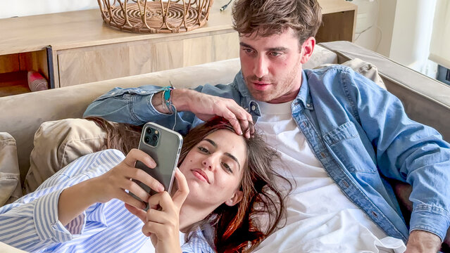 Young couple lying on a comfortable sofa, the woman holding a smartphone while the man looks over her shoulder, sharing an online experience at home