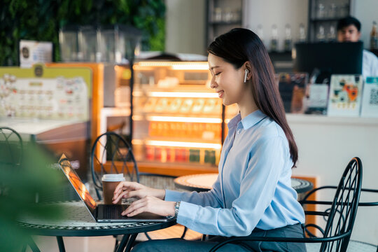 focused Asian businesswoman typing on laptop computer at cafe with coffee paper cup on table