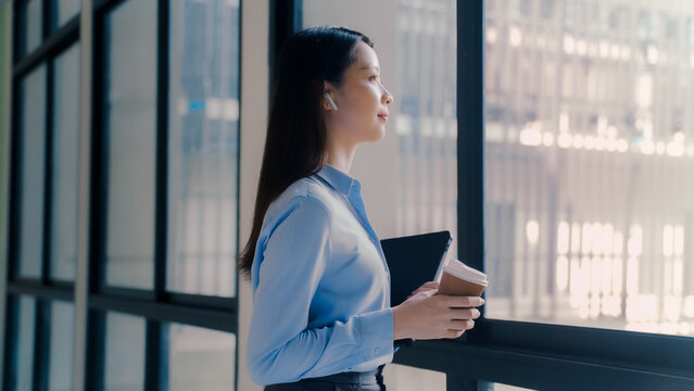 Thoughtful Asian businesswoman holding coffee cup looking out window with sunlight