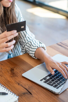 Woman holding a credit card while typing on a laptop, making an online purchase and performing e-commerce transactions on a wooden table