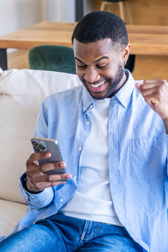 Smiling young black man with a cheerful expression raising his fist in triumph while looking at his mobile phone, celebrating good news or an online win at home on the sofa