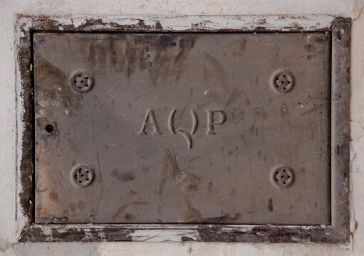 Close-up of a weathered metal utility panel with an AQP inscription (Acquedotto Pugliese), set into a distressed wall. Features a grunge aesthetic and industrial details.
