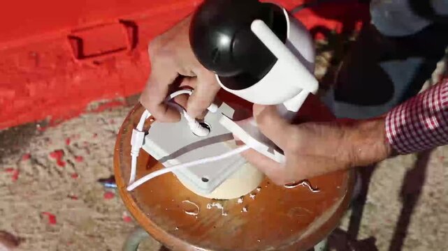 Professional electrician inserts a CCTV security camera cable into a junction box before mounting it on an exterior house wall during surveillance system installation.