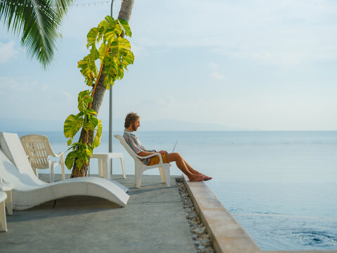 Man working with laptop sitting by the pool at seaside 