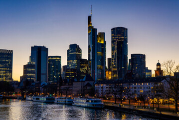Fototapeta premium Frankfurt, Germany, a view of the illuminated financial district from the Main river with sightseeing boats at blue hour and cloudless sky in the evening
