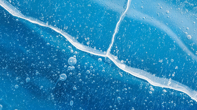 Macro photography of frozen natural ice texture with trapped air bubbles and large structural fractures creating beautiful geometric patterns in deep blue color tones.