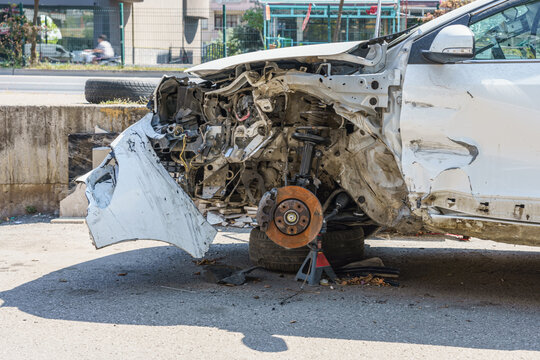 Damaged white car with exposed engine components and missing front wheel resting on a tire, parked on a concrete surface near a busy street