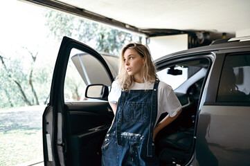 Woman in overalls stands near car in garage during daytime © Bohdan