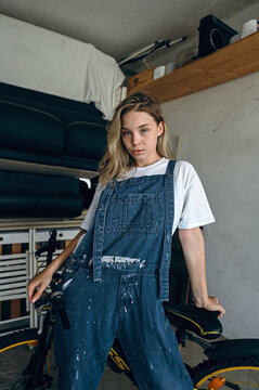 Young woman stands beside bicycle in a garage while wearing denim overalls and a simple white shirt