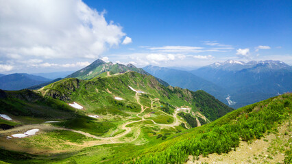 Fototapeta premium Wide landscape view of a green mountain range in summer, Rosa Khutor resort, Sochi, Russia. Winding dirt road and trails on alpine hills with patches of snow. Blue sky over distant peaks