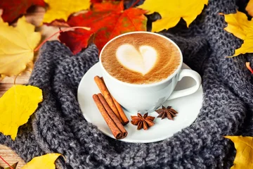 Cozy autumn still life featuring a cup of creamy cappuccino with heart latte art, cinnamon sticks and colorful fall leaves on a knitted blanket. © preto_perola