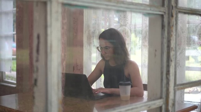 Young woman working on laptop and drinking coffee at table on old glass veranda