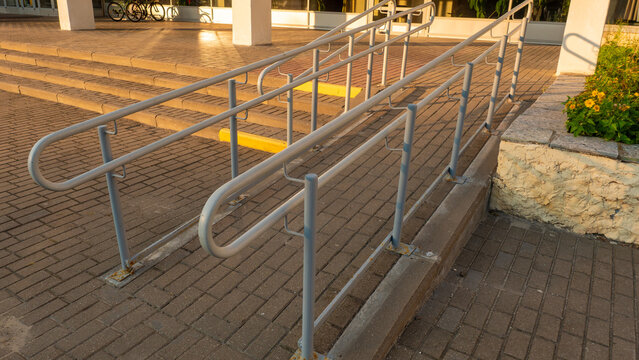 Angled perspective of concrete wheelchair ramp featuring double metal handrails installed on stone tile pavement