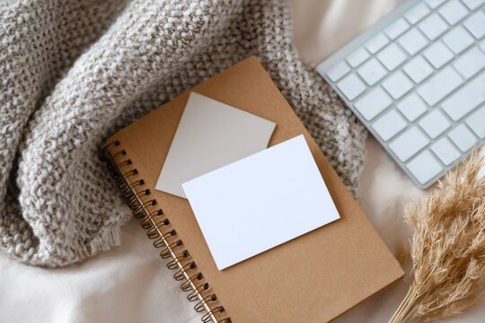 Cozy home office flat lay with branding mockup. Blank business cards on a notebook with a knit blanket and keyboard. Neutral hygge workspace concept