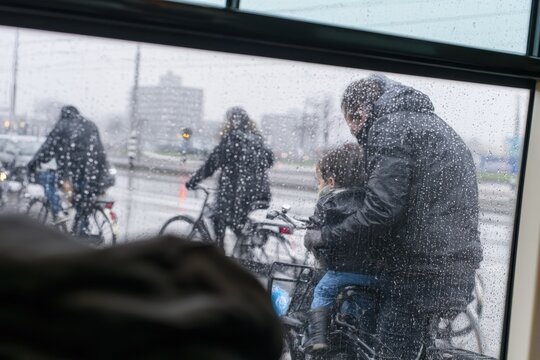people bundled in winter coats ride bicycles in the netherlands during a light snowfall as seen from a window