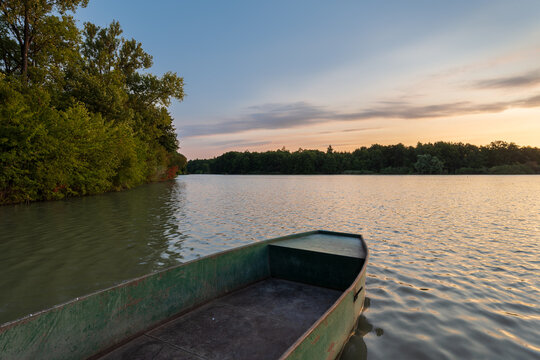 Old green metal rowboat anchored on a fish breeding pond at sunset. Scenic countryside landscape with a fishpond surrounded by lush forest under a warm twilight sky.