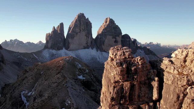 Dynamic drone shot flying toward Tre Cime di Lavaredo close past cliffs at sunrise