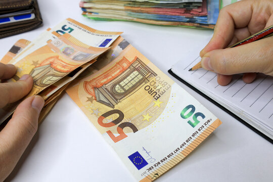Hands counting &euro;50 euro bills while writing on a notepad, with a stack of mixed euro denominations in the background